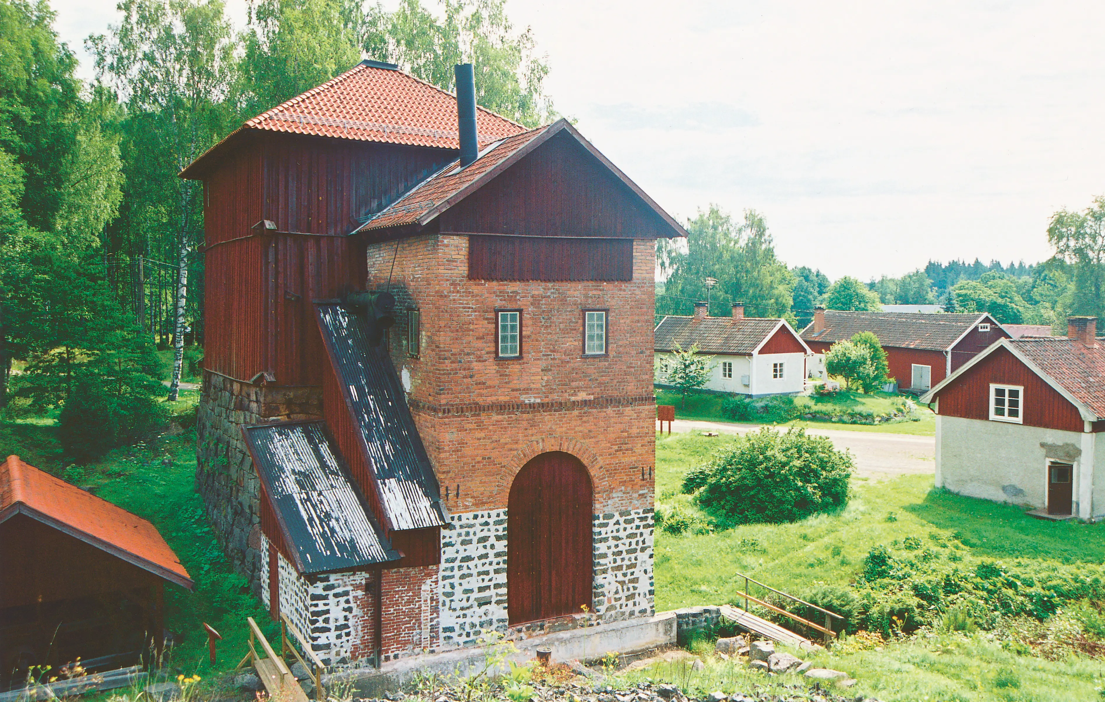 Igelbäckens masugn. En hög, smal byggnad i sten, tegel och rödmålat trä. Sommar och grönska.
