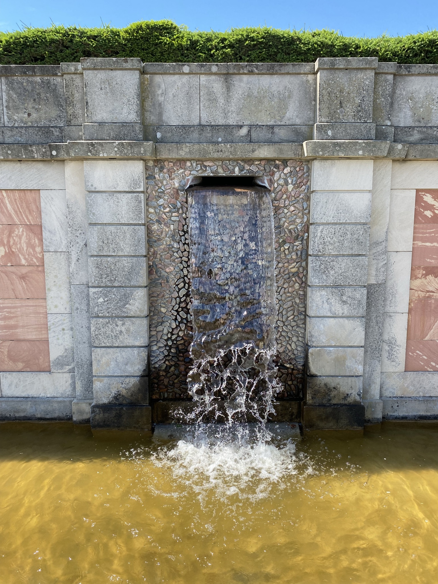 Drottningholms slottspark. En mur av olika sorters sten, med en öppning där vatten strömmar ut och faller ned i en grund bassäng.