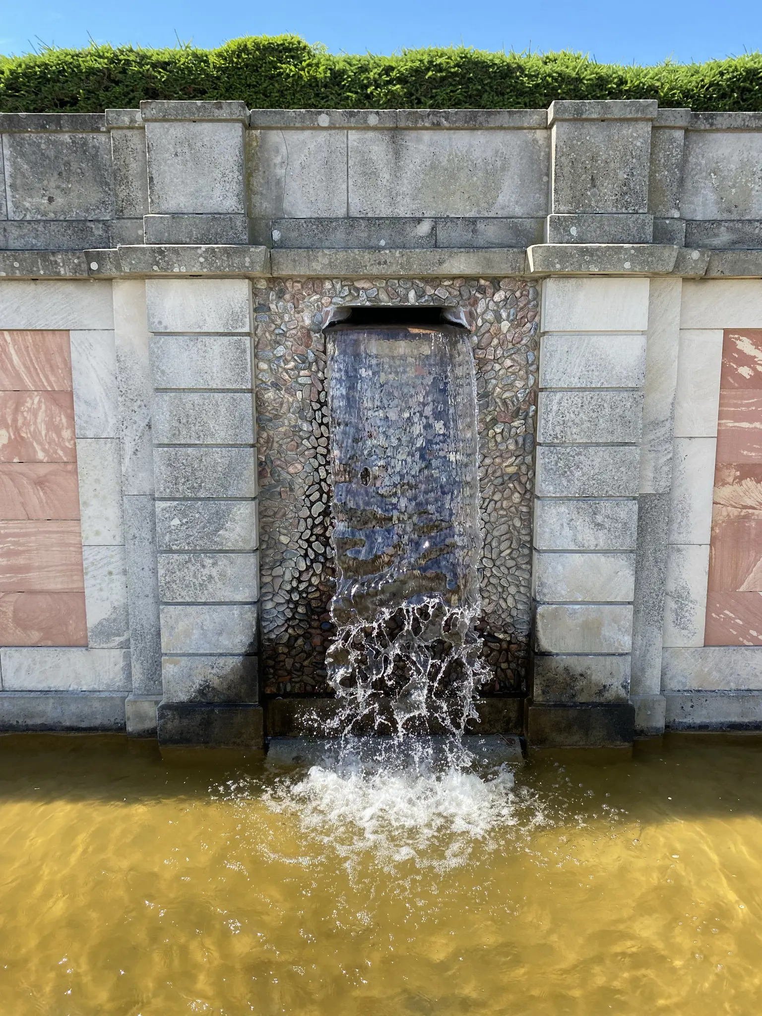 Drottningholms slottspark. En mur av olika sorters sten, med en öppning där vatten strömmar ut och faller ned i en grund bassäng.