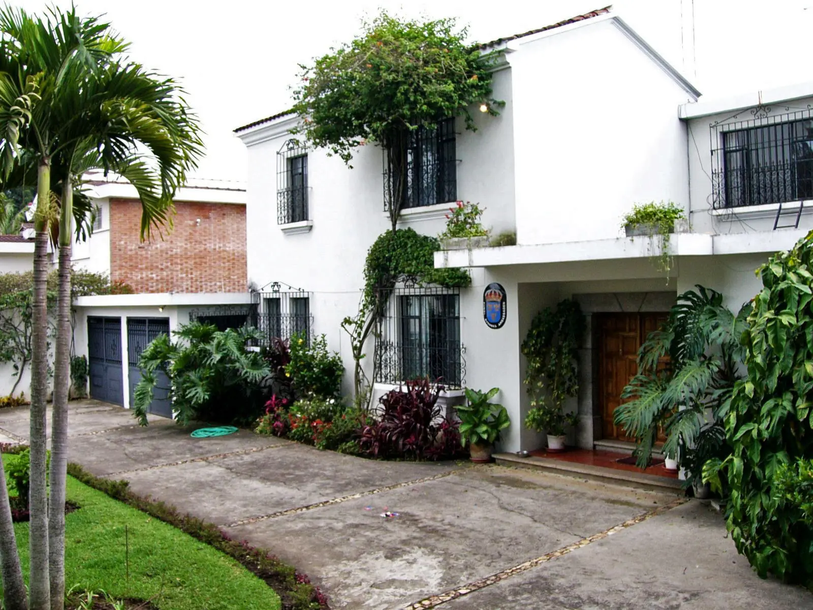 Chefsbostad, Guatemala City. En vitputsad tvåvåningsbyggnad med svarta smidesgaller över fönstren, omgiven av palmer och planteringar.