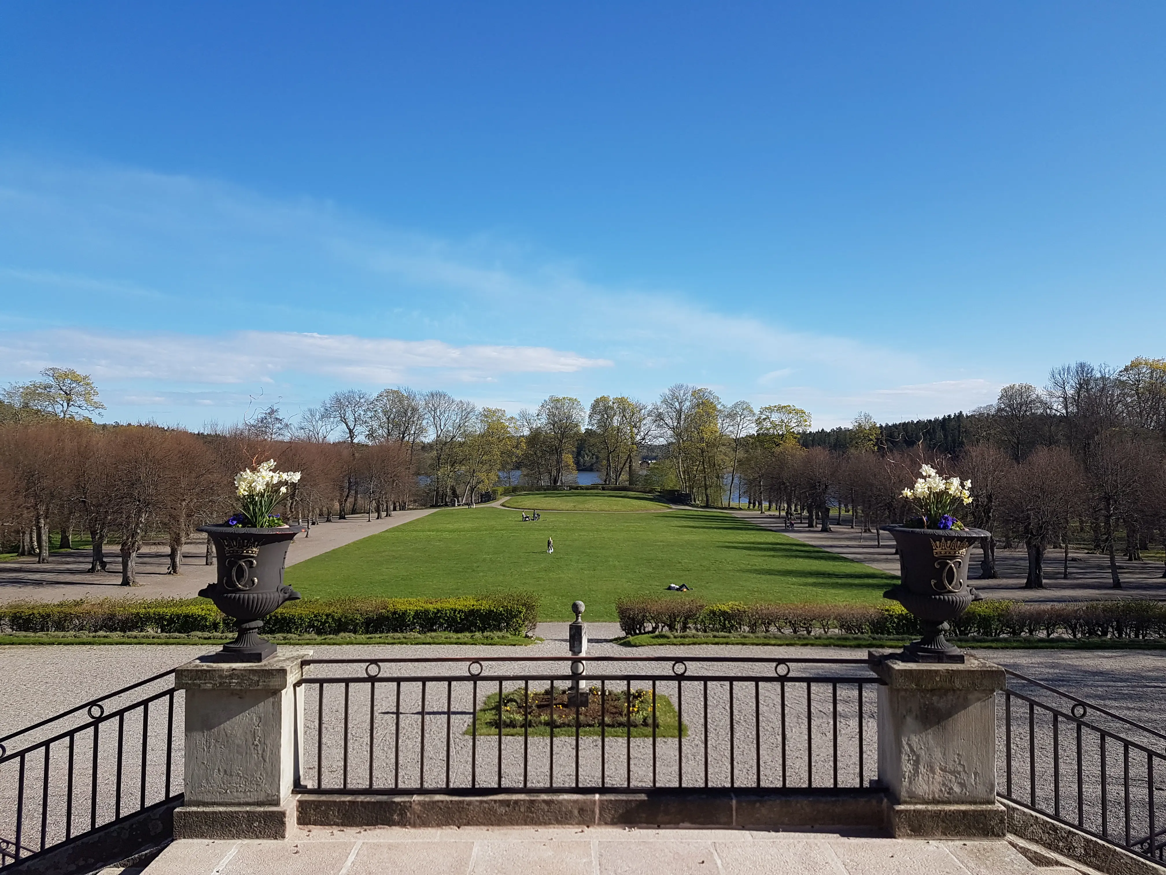 Vy från Hoglandsterassen på Rosersbergs slott. Mot vattnet sträcker sig en gräsmatta där folk solar och promenerar. Vår och grönska.