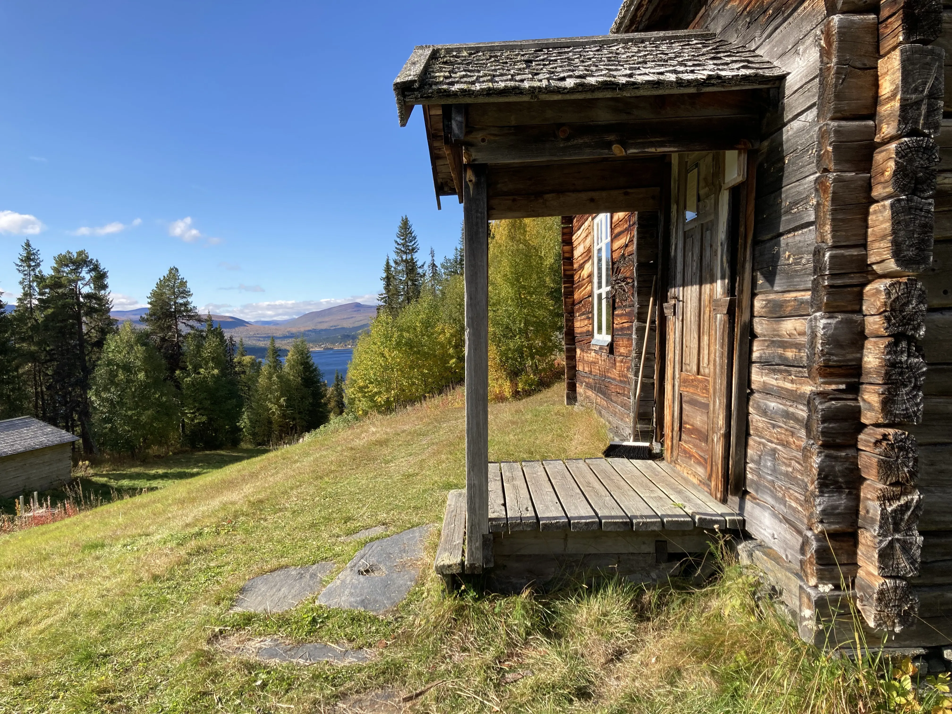 Till höger i bild syns en farstukvist till en gammal timmerstuga, till vänster i bild skymtar ett vidsträckt fjällandskap och en sjö.