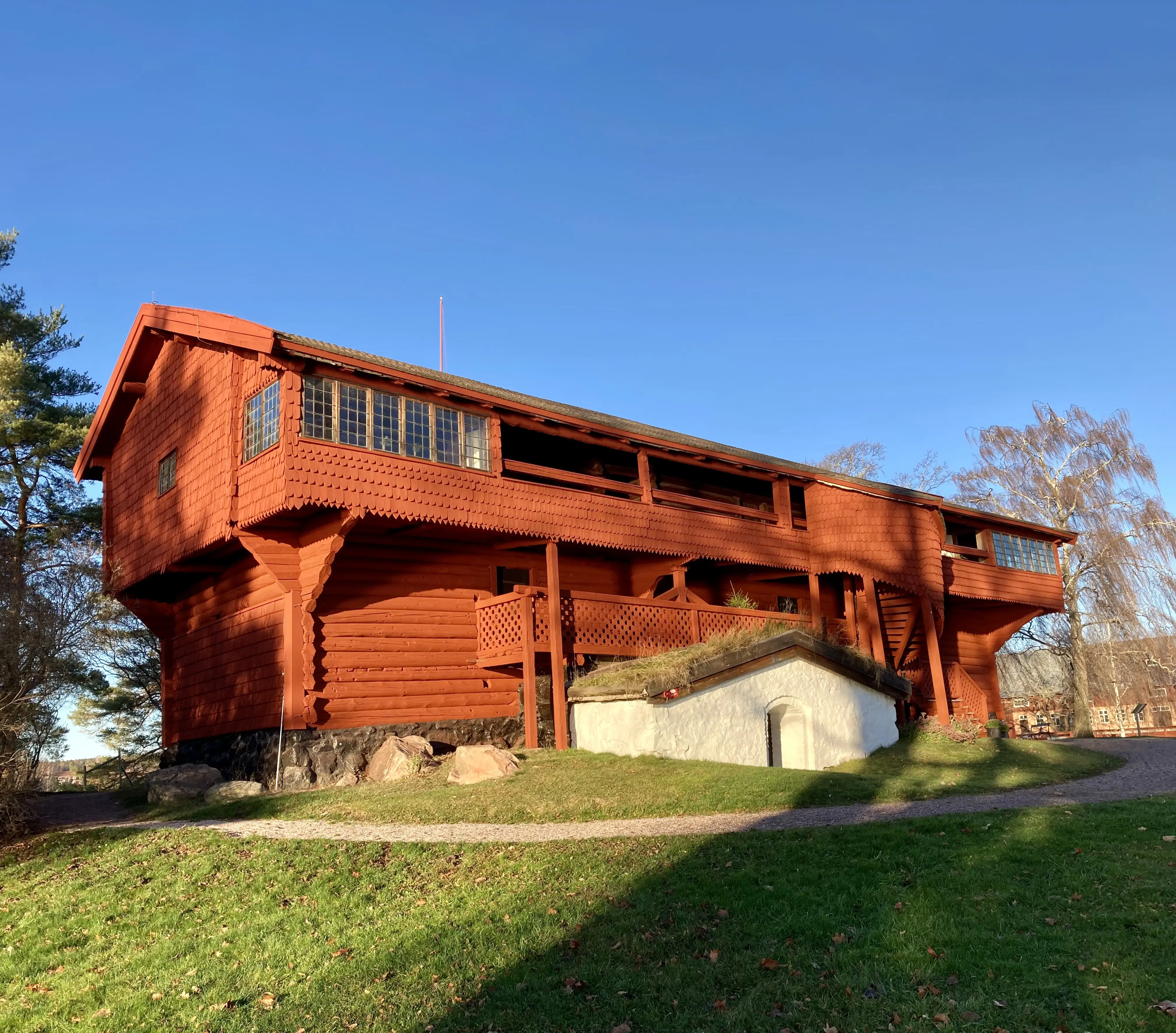 Ornäs-stugan med solbelyst, klar-röd fasad. Huset är byggt i liggande timmer och har ett utskjutande loft, klätt med träspån.