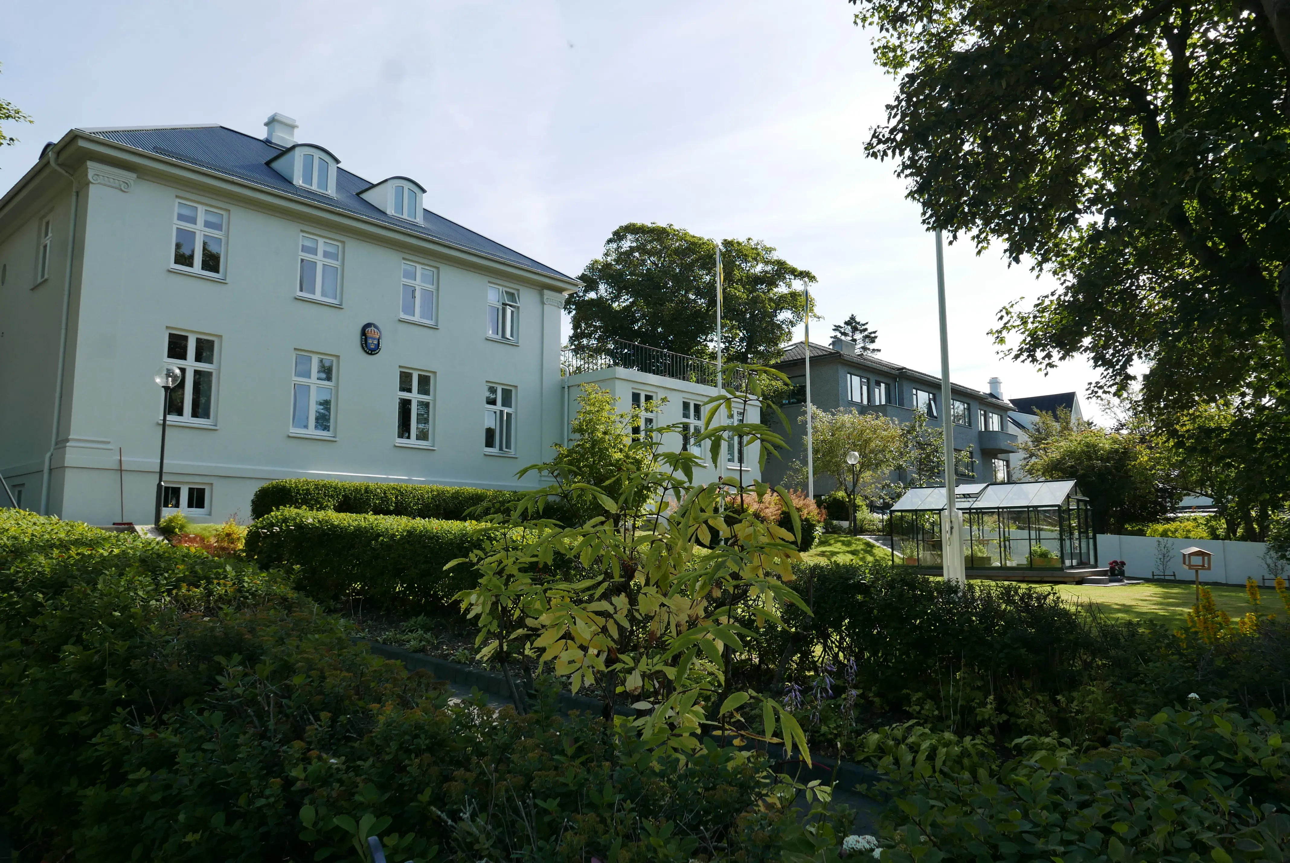 Reykjavik, ambassadörsbostaden. En stor, vitputsad villa omgiven av en lummig trädgård. Sommar och grönska.