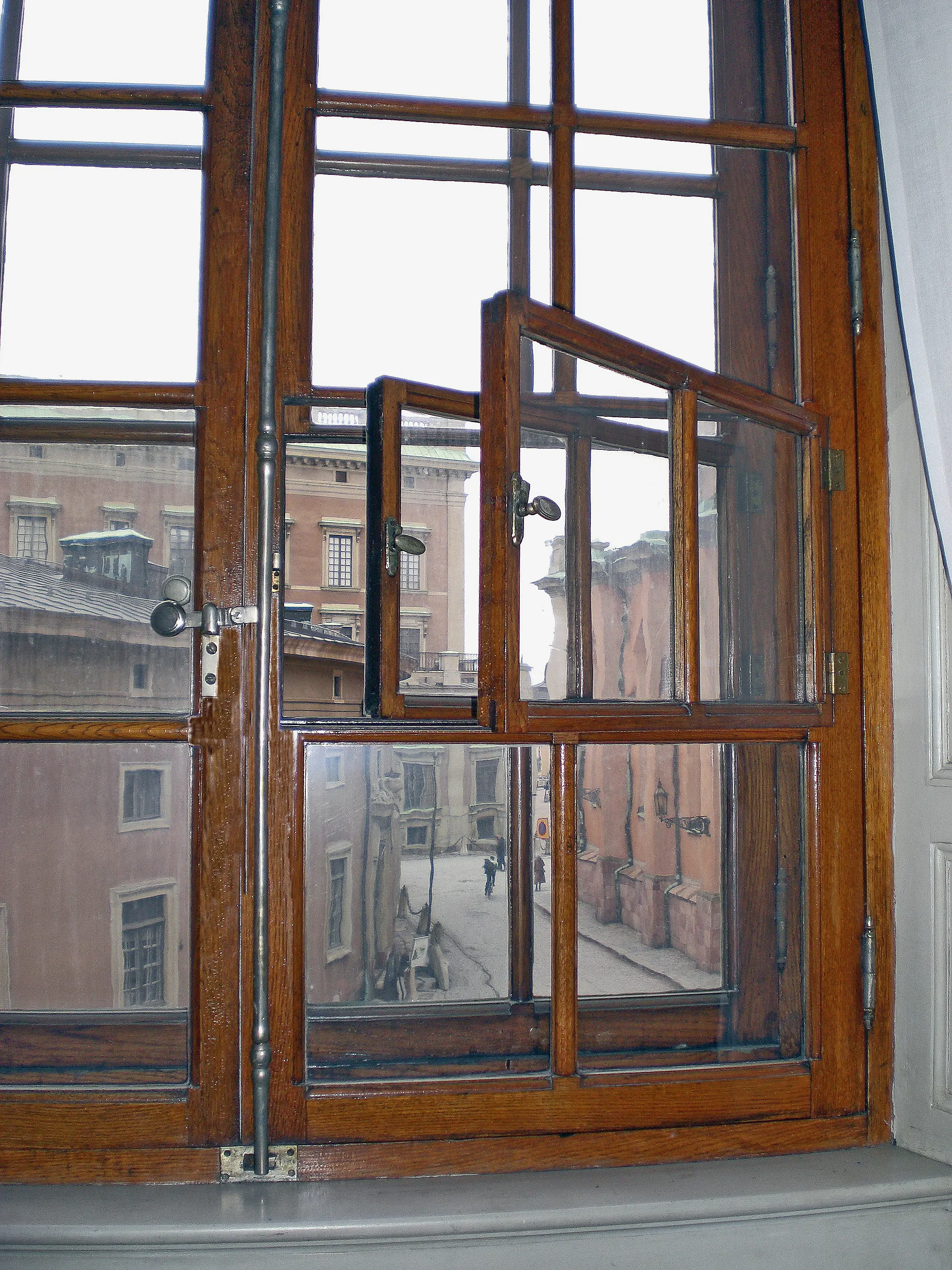 Ett spröjsat fönster med mörk träram och en liten öppningsbar ruta mitt i fönstret. Fotot är taget inifrån och ut genom fönstret är utsikt mot stadsbyggnader.