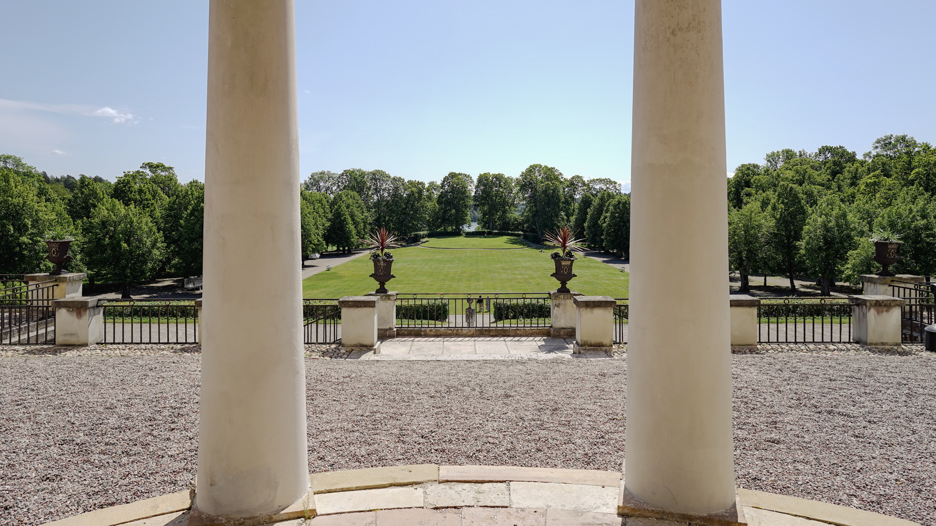 Rosersbergs slott. Vy från Hoglandsterassen och en entré med pelare. Mot vattnet sträcker sig en gräsmatta. 
