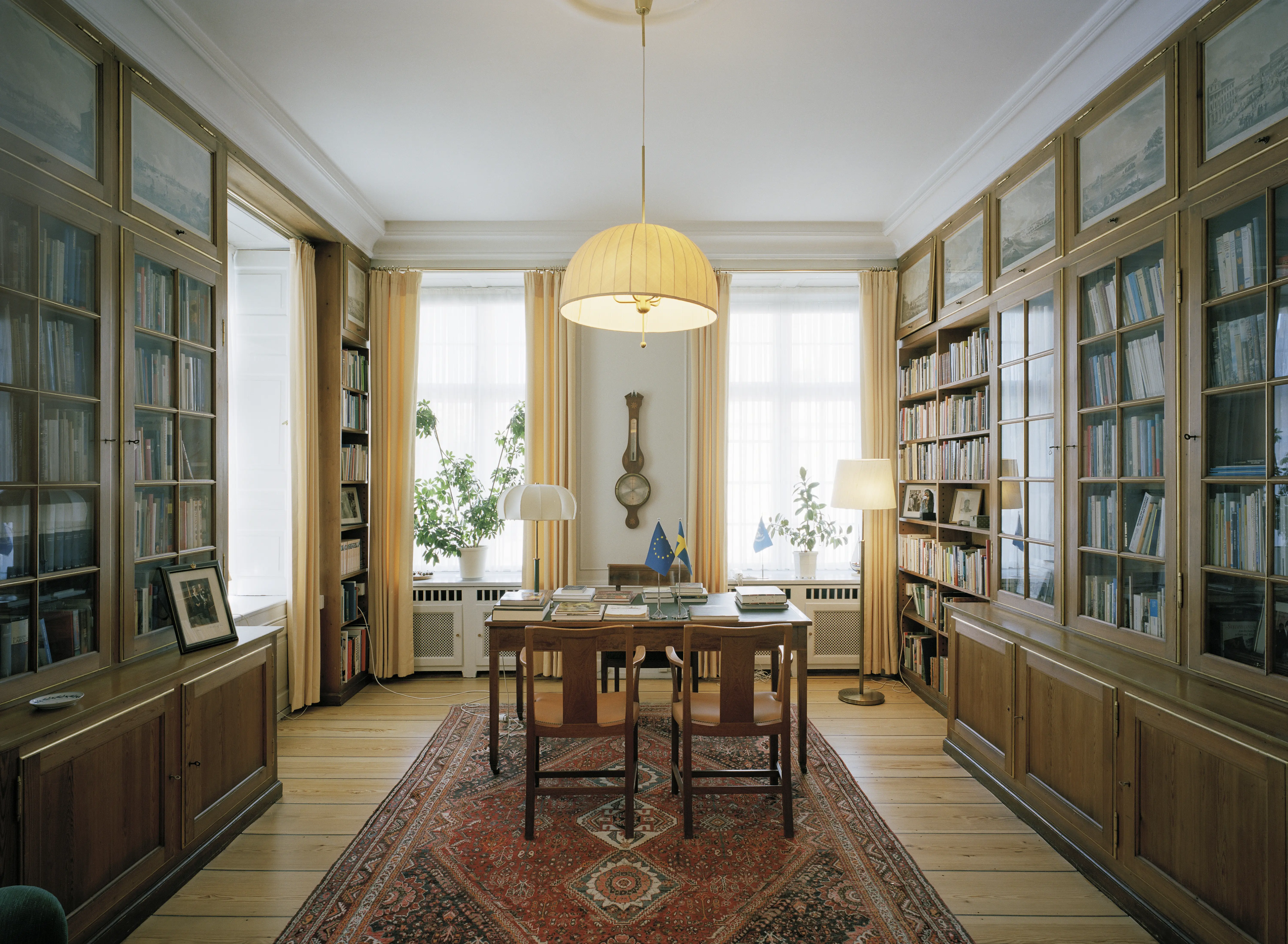 Ambassaden i Köpenhamn, interiör. Bibliotek med öppna hyllor och vitrinskåp. Stora fönster och lampor med ljusa skärmar ger ett milt ljus.