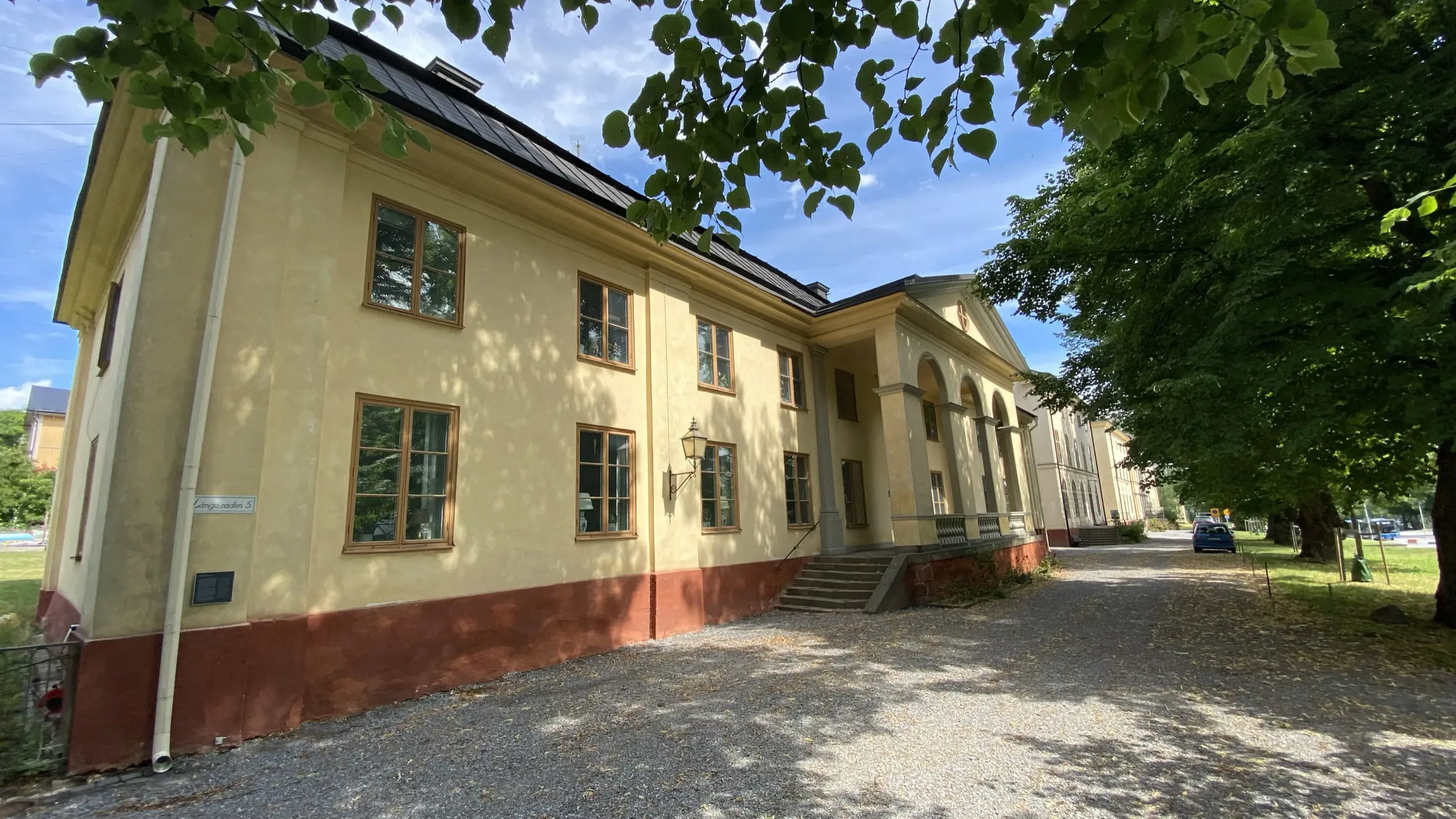 Kanslihuset på Malmen vid Drottningholm. Gulputsad tvåvåningsbyggnad med röd sockel och pampig portik. Sommar och grönska.