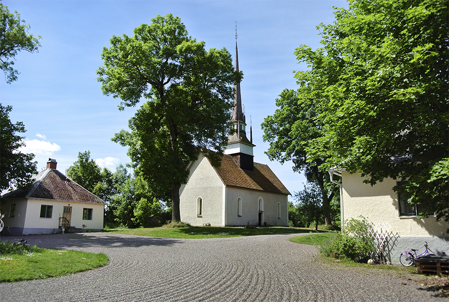 En liten kantig ljusputsad kyrka med ett torn och spira. En krattad grusväg syns i förgrunden och kyrkan omges av en blå himmel och grönskande träd.