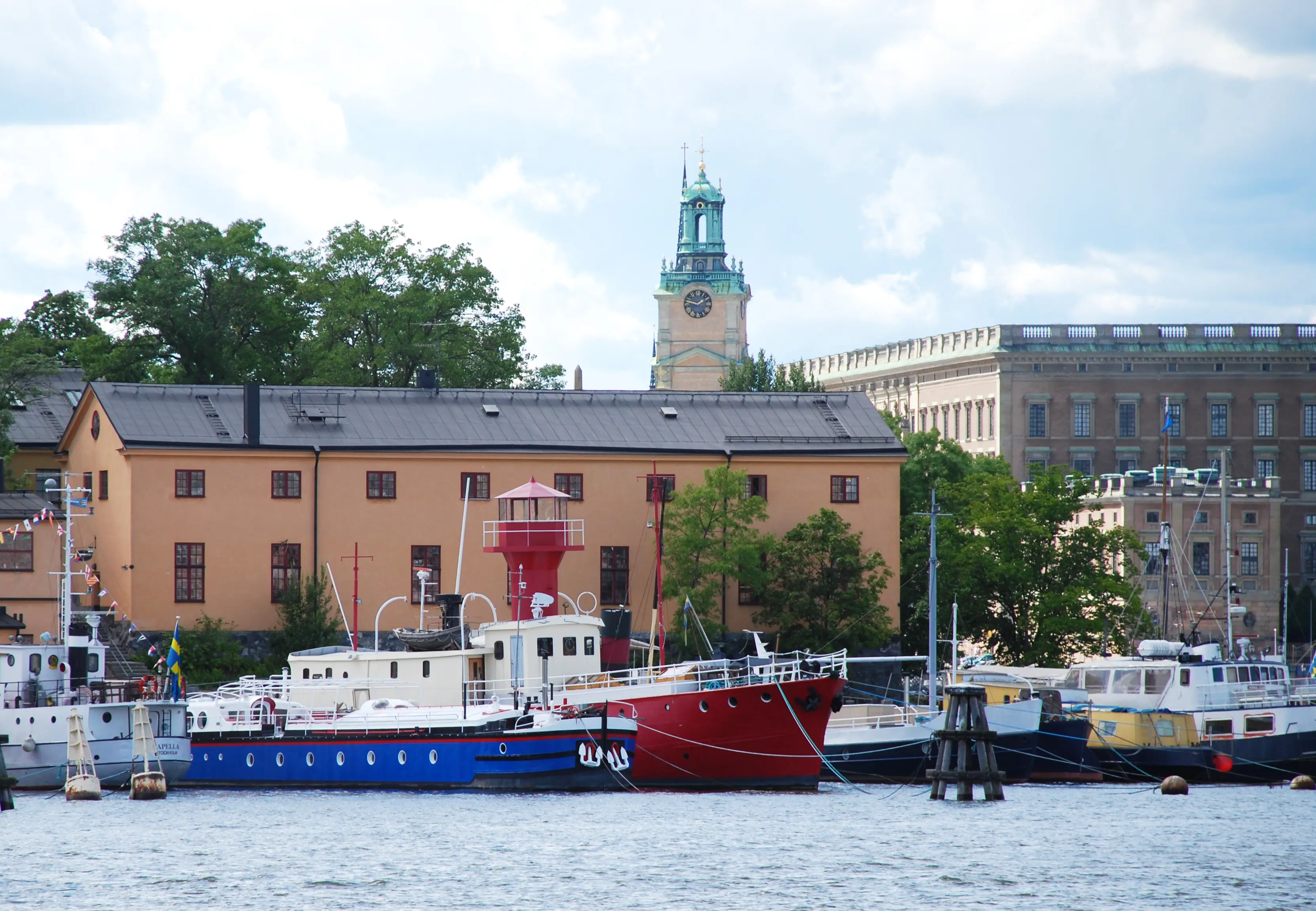 Råseglarhuset på Skeppsholmen. En enkel, gulputsad tvåvåningsbyggnad vid en kaj med stora båtar. I bakgrunden syns Stockholms slott.
