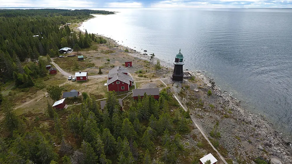 Flygfoto av Berguddens fyrplats. Fyrtornet ligger vid den steniga stranden. Rödmålade hus är omgivna av barrskog. Vy ut mot Kvarken.