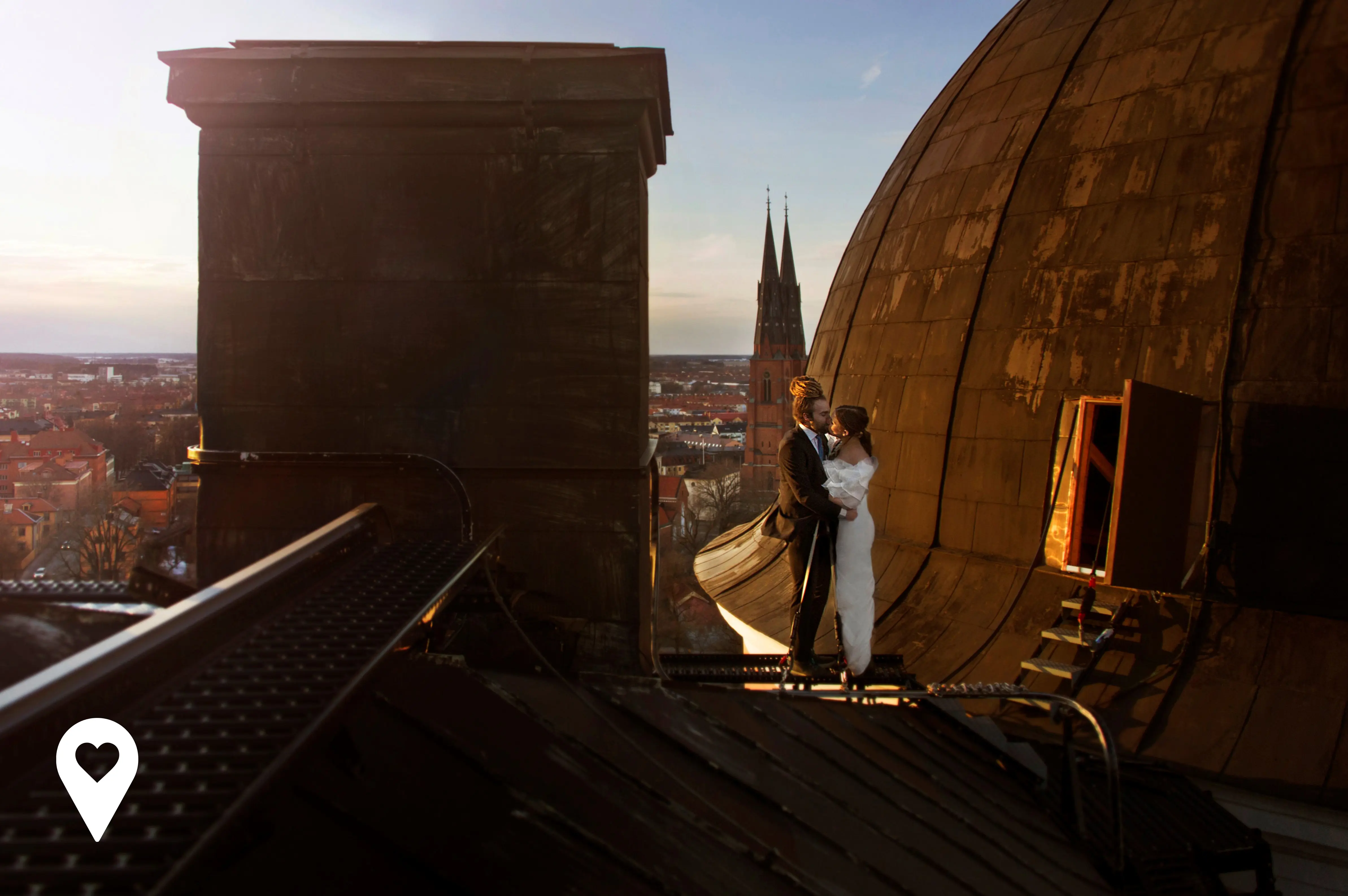 Ett par i bröllopskläder håller om varandra uppe på taket på Uppsala slott. Storslagen vy av Uppsala i bakgrunden