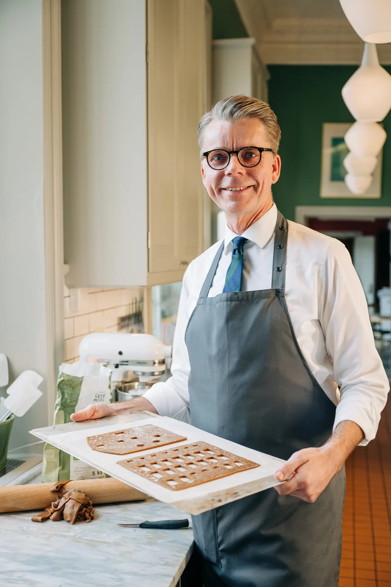 En man i vit skjorta, svarta glasögonbågar och ett grått förkläde står med ett stort leende och håller i en plåt med utkavlad pepparkaksdeg som han skurit ut till husdelar till ett pepparkakshus.