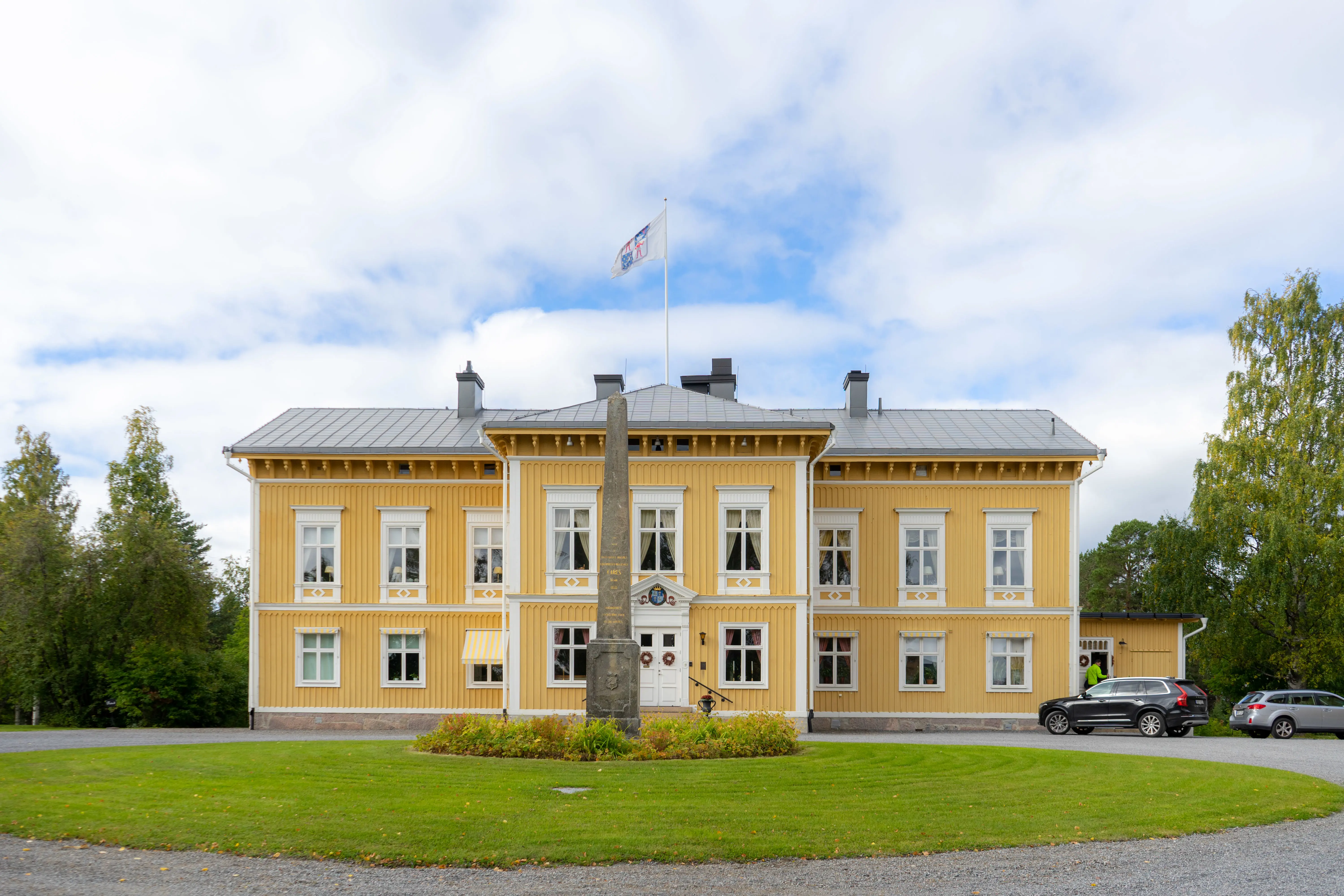 Residenset i Luleå. En gul träbyggnad i två våningar, med vita snickerier. Framför residenset står en stenobelisk. Höst.