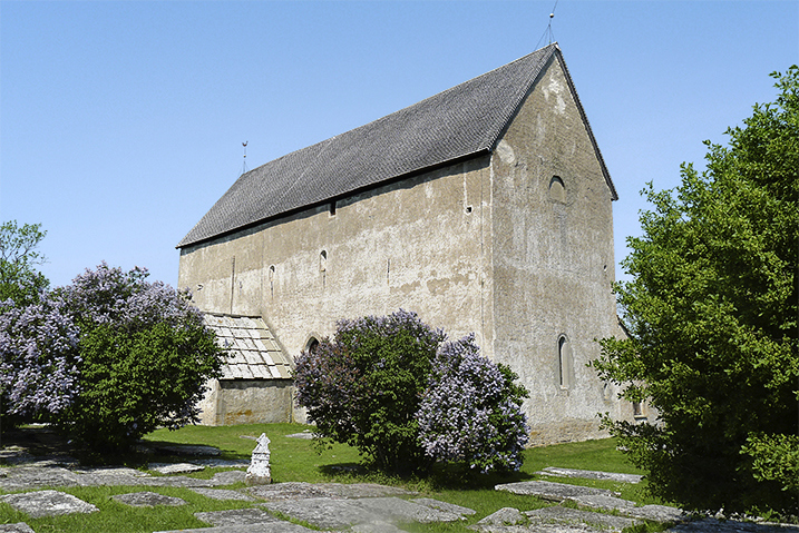 En kantig hög kyrka med en grå ton på fasaden. Ett sluttande tak i mörkt gråbrunt. Kyrkan omges av grönska och blommande syrénbuskar.