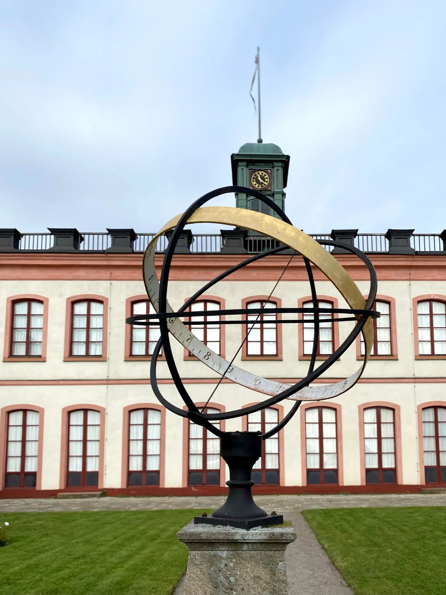 Ett solur i förgrunden och en stor rosa och ljus byggnad med ett ur på taket.