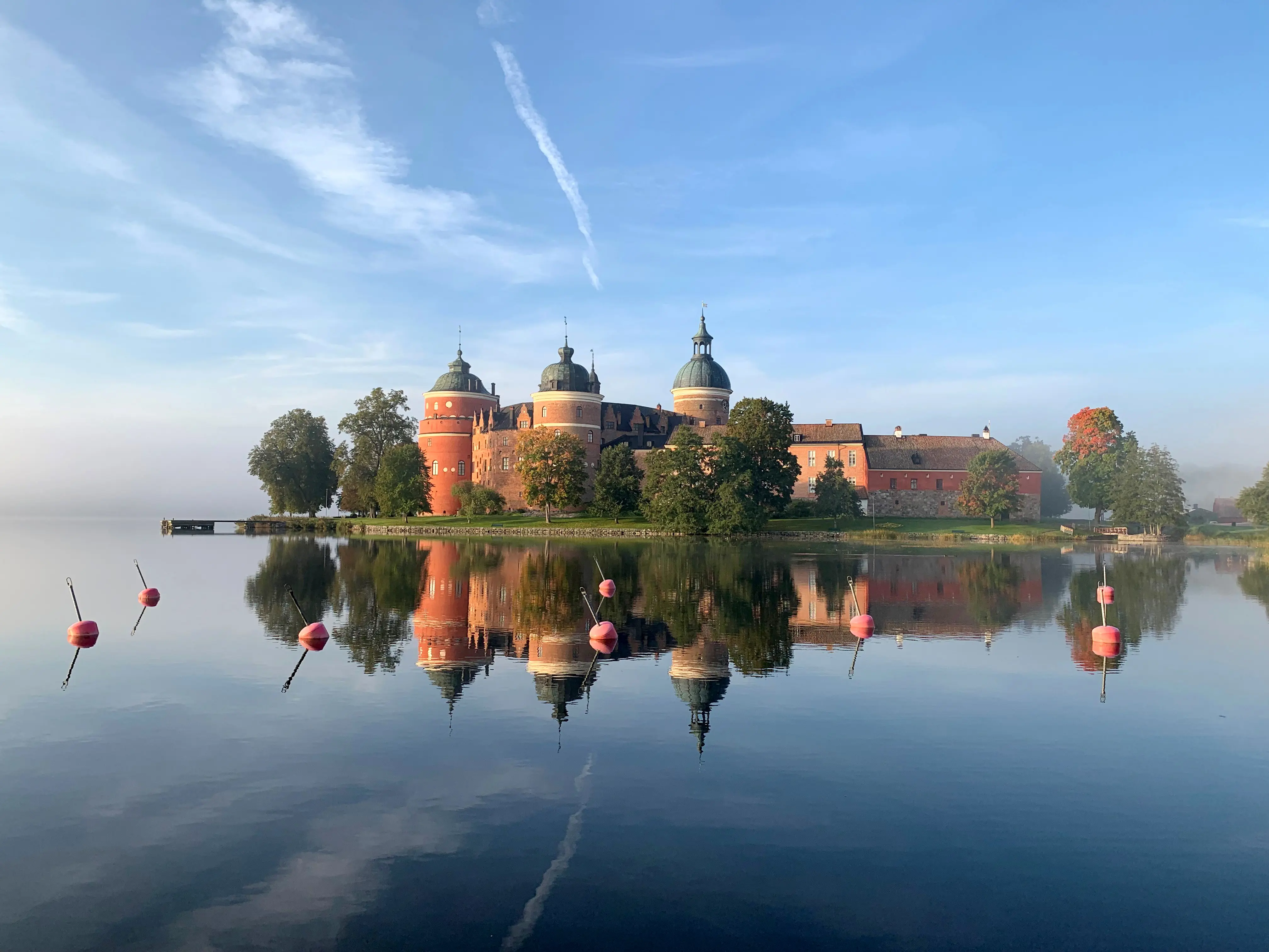 Tegelrött slott vid spegelblankt vatten med runda torn, insvept i grönska.