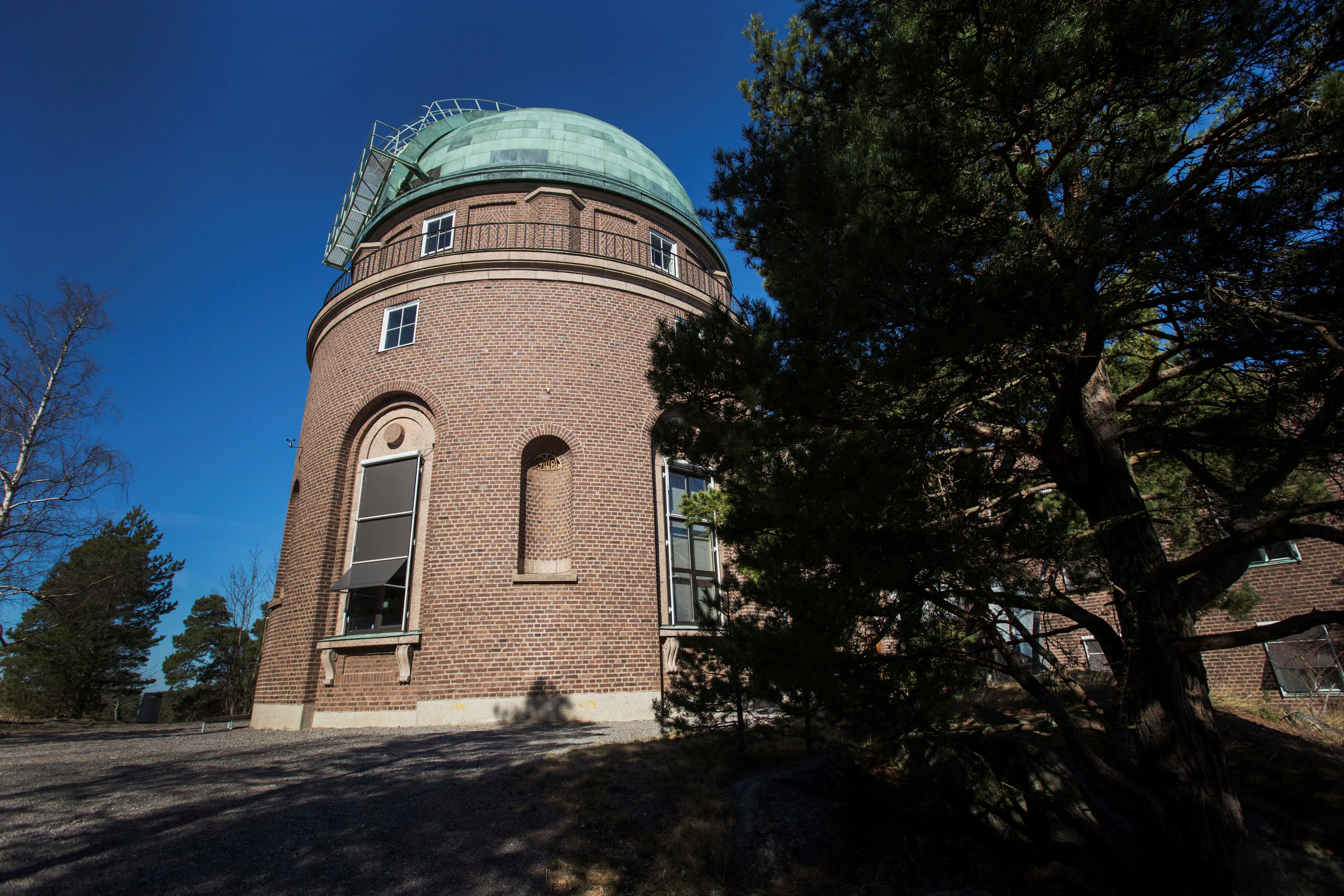 Teleskopbyggnaden vid Stockholms observatorium. Ett runt tegeltorn med kopparkupol, omgivet av tallar.