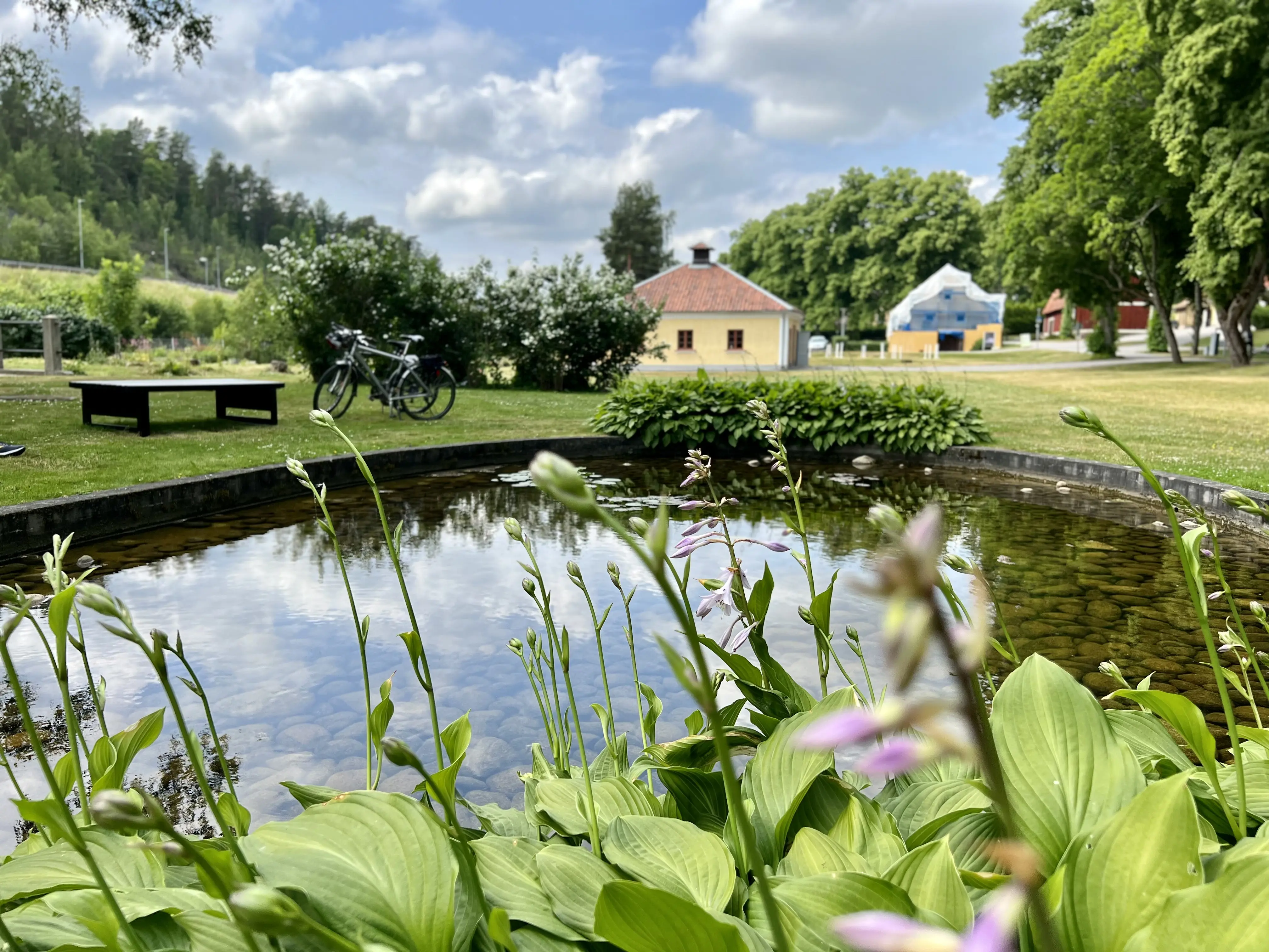 Tumba bruk. En liten damm med sittbänk och blommor. I bakgrunden skymtar spruthuset. Sommar och grönska.