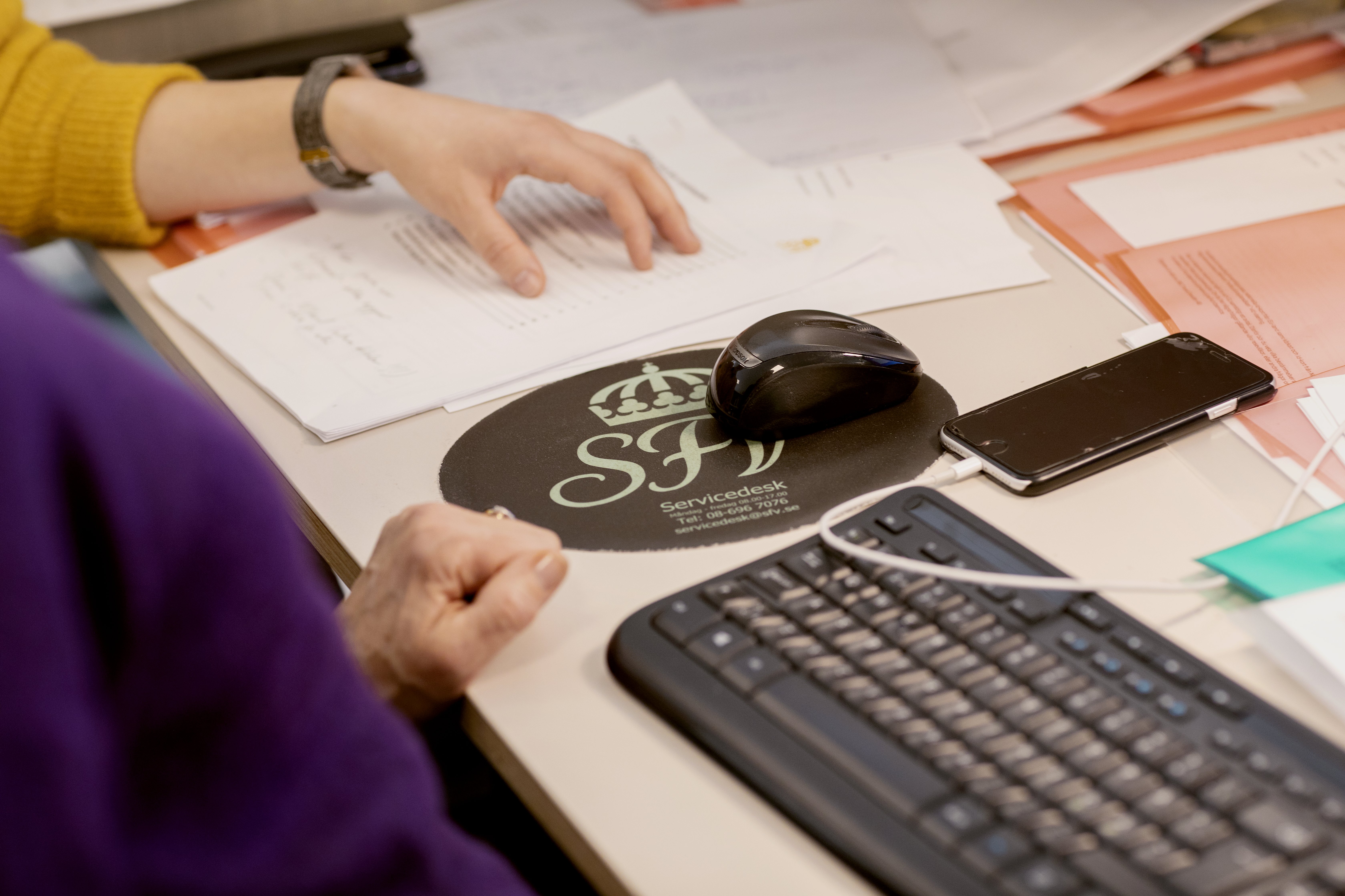 Närbild på kontorsplats med tangentbord, datormus, mobiltelefon och dokument. Två personers händer syns.