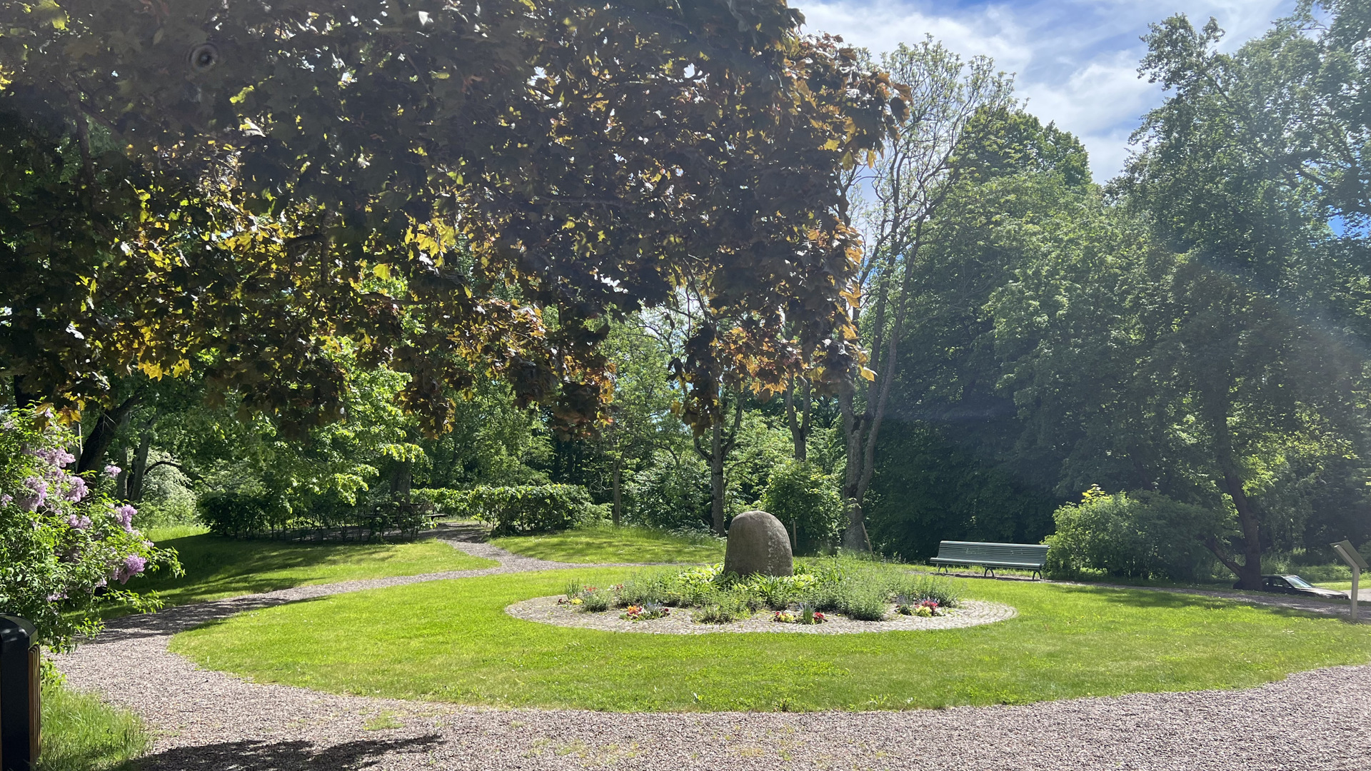 Bogesunds slottsträdgård. Brahestenen, inramad av blomsterplanteringar, gräsytor, grusgångar och sittbänkar. Sommar och grönska.