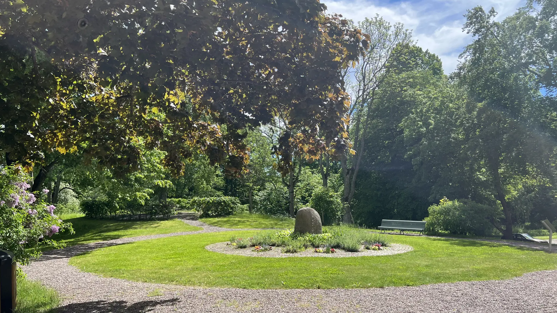 Bogesunds slottsträdgård. Brahestenen, inramad av blomsterplanteringar, gräsytor, grusgångar och sittbänkar. Sommar och grönska.