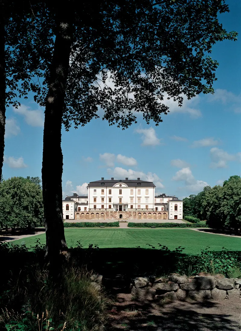 Rosersbergs slott med sin vitputsade fasad sett på avstånd med ett lövträd i förgrunden.