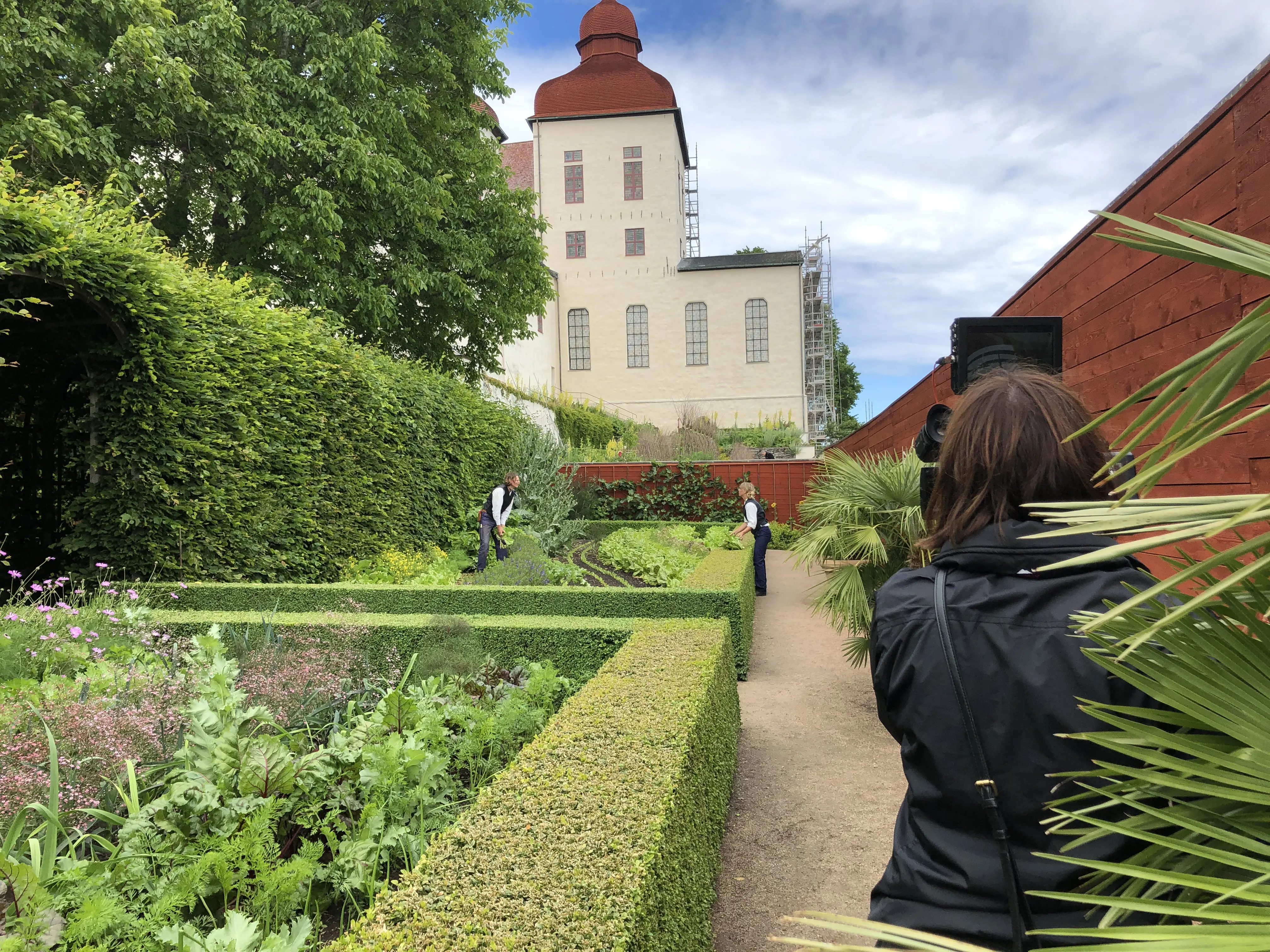 En person står framför en kamera och filmar två andra personer i en trädgård.