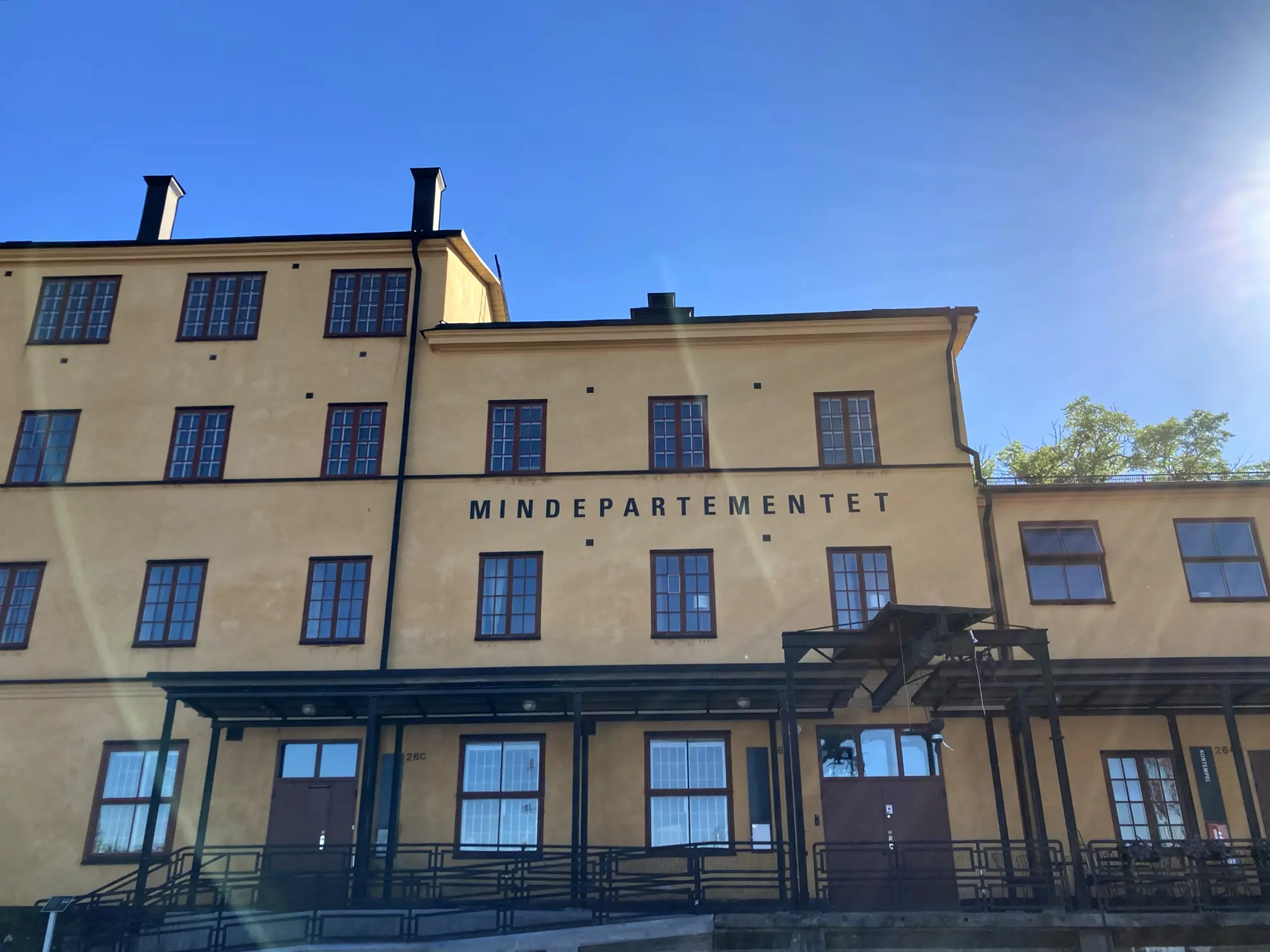 Mindepartementet, Skeppsholmen. En gulputsad tvåvåningsbyggnad med stora fönster och plåtdörrar. Sommar och grönska.