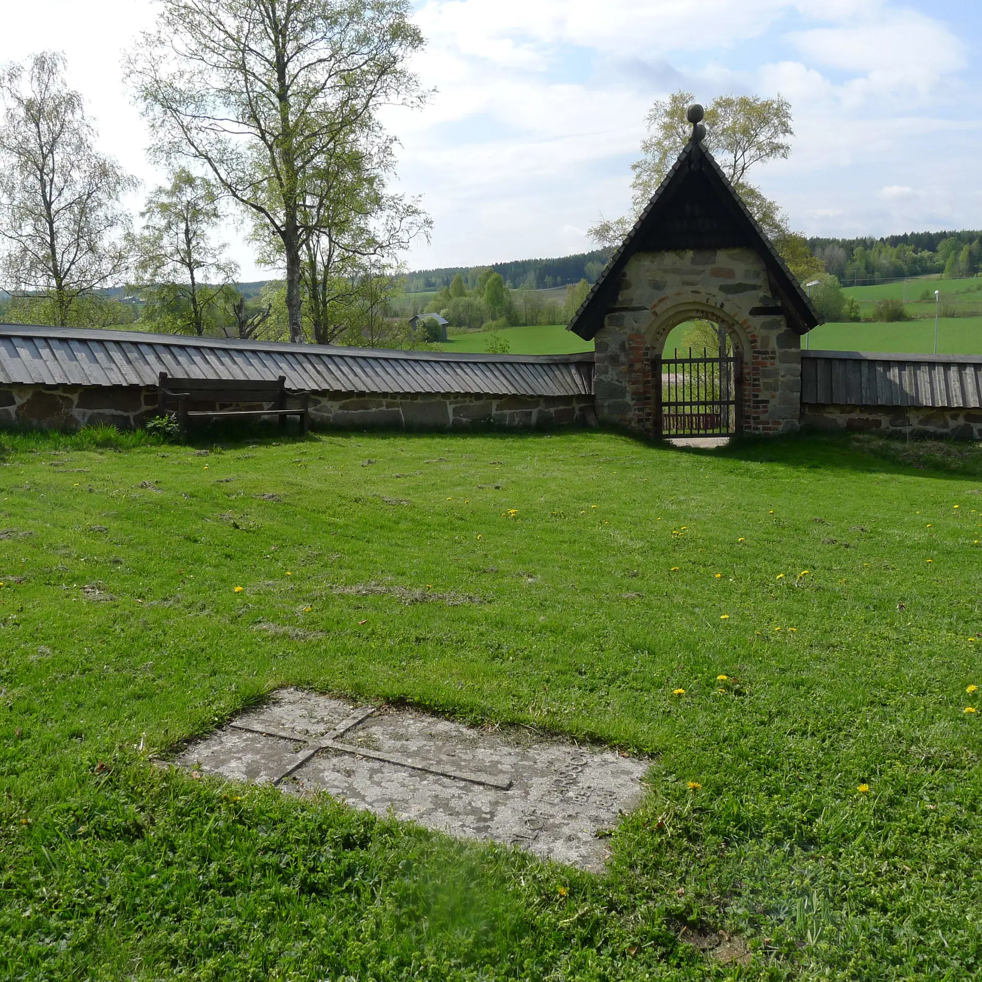 Trönö gamla kyrkas kyrkogård. I gräset vilar en gravhäll. Bortanför muren och dess portvalv syns ett kuperat jordbrukslandskap.