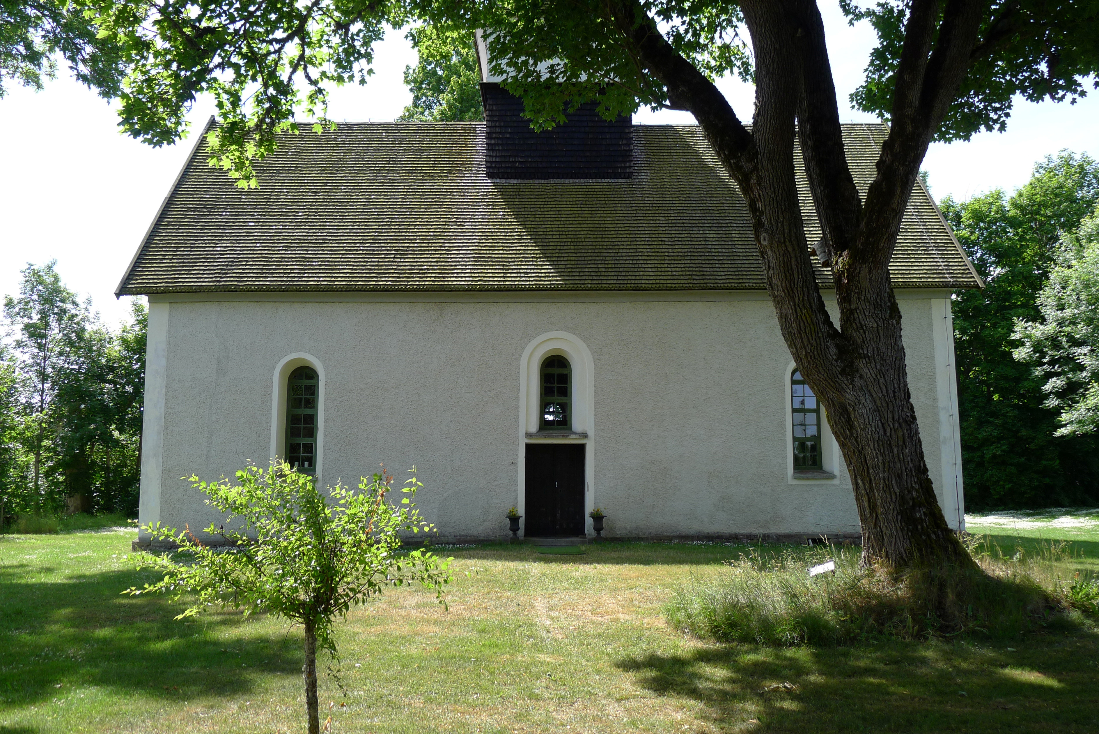 Brunneby kyrka. En liten, vitputsad medeltida kyrka med rundbågiga fönster. Sommar och grönska.