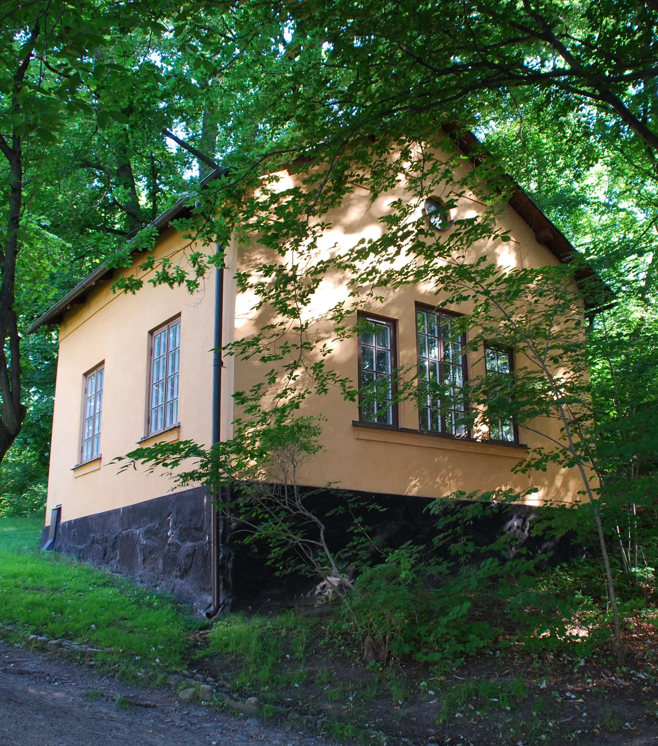 Gamla Skogsinstitutets tvättstuga i gulputsad sten. Huset står i en backe. Sommar och grönska.