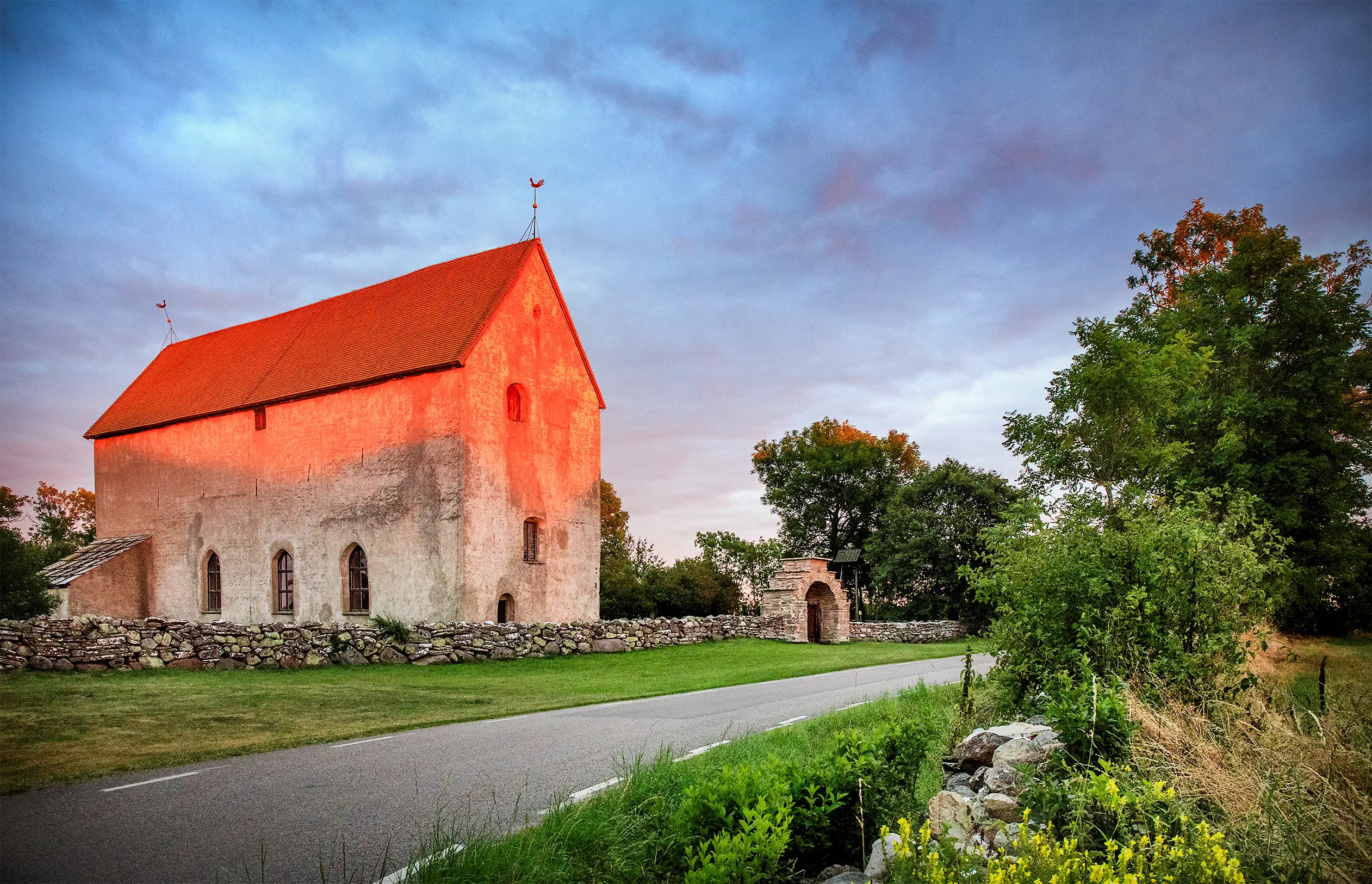 Källa gamla kyrka i kvällsljus. Den medeltida stenfasaden skimrar röd. En smal landsväg går längs stenmuren med sin trappstegsformade port.