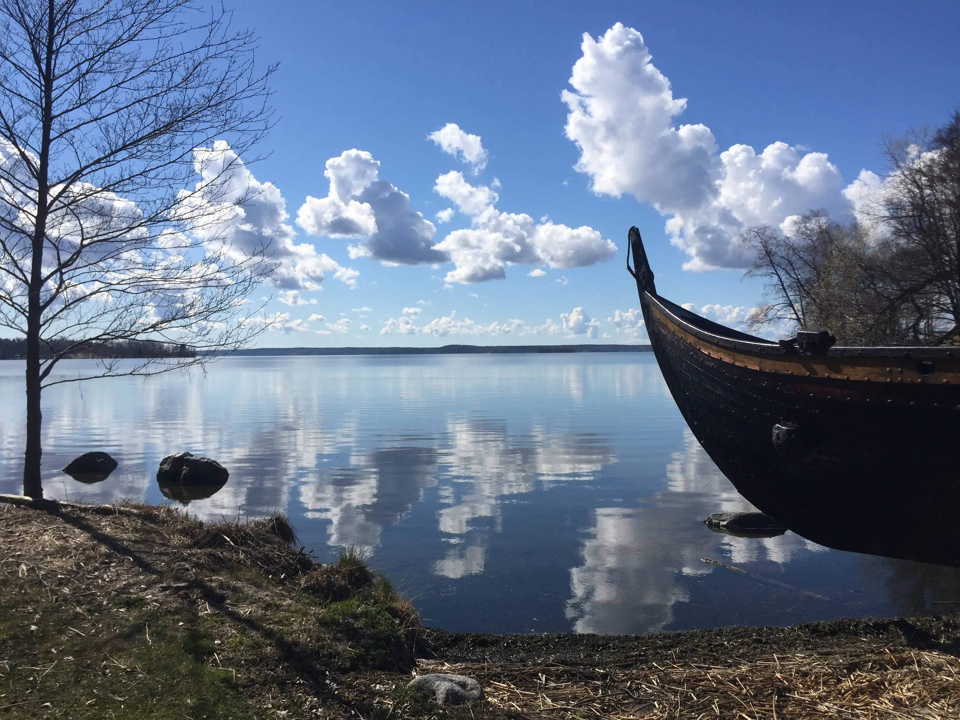 En del av en båt av vikingastil vid en sjö. Molnen på en klarblå himmel speglar sig i vattnet.