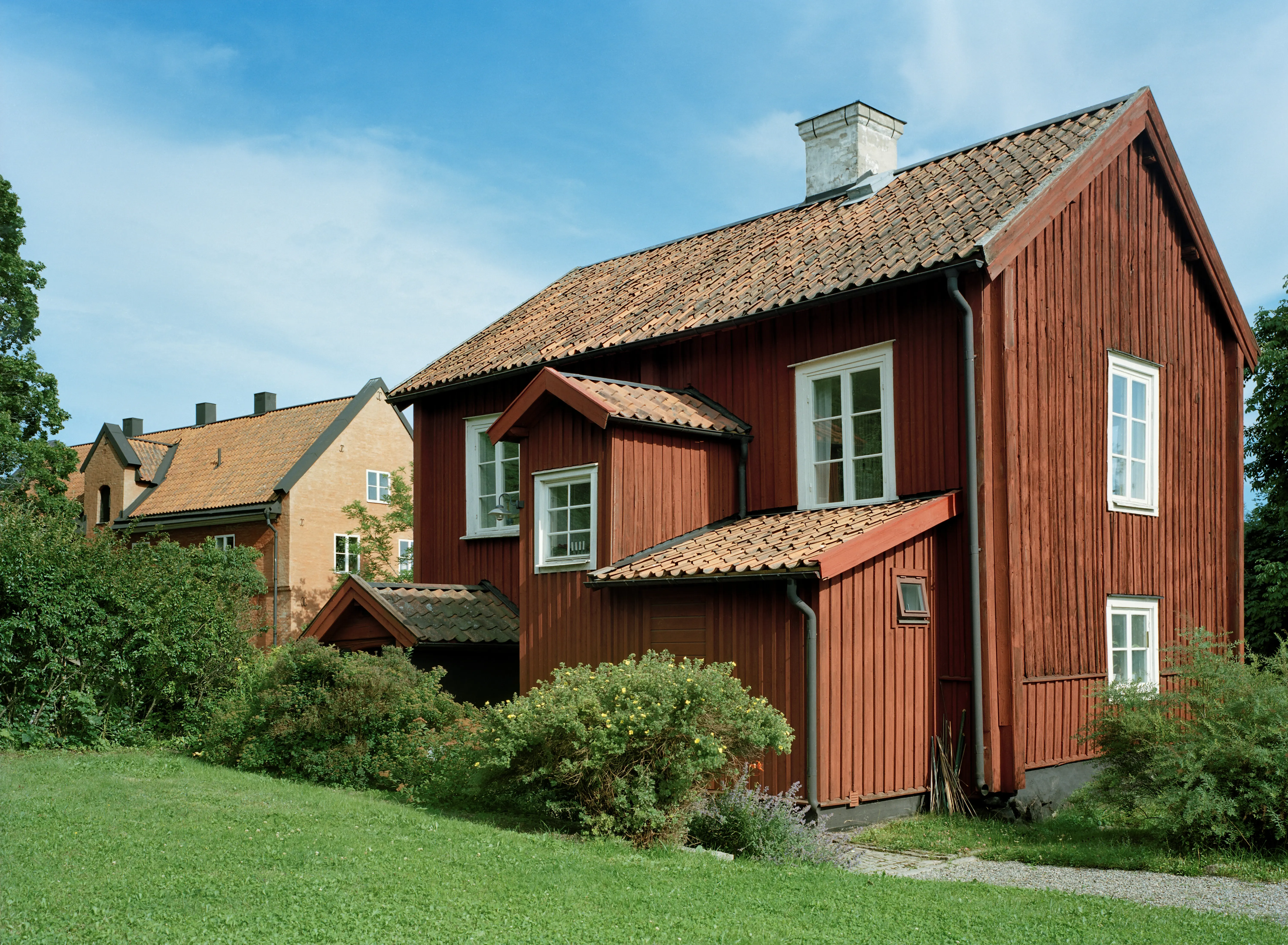 Gamla biskopsgården. Tvåvåningshus klätt i stående, rödmålad träpanel. Trapphus och förråd är påbyggda. Sommar och grönska,